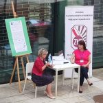 Dwie kobiety czytające książkę, siedzące na krzesłach przy stoliku.